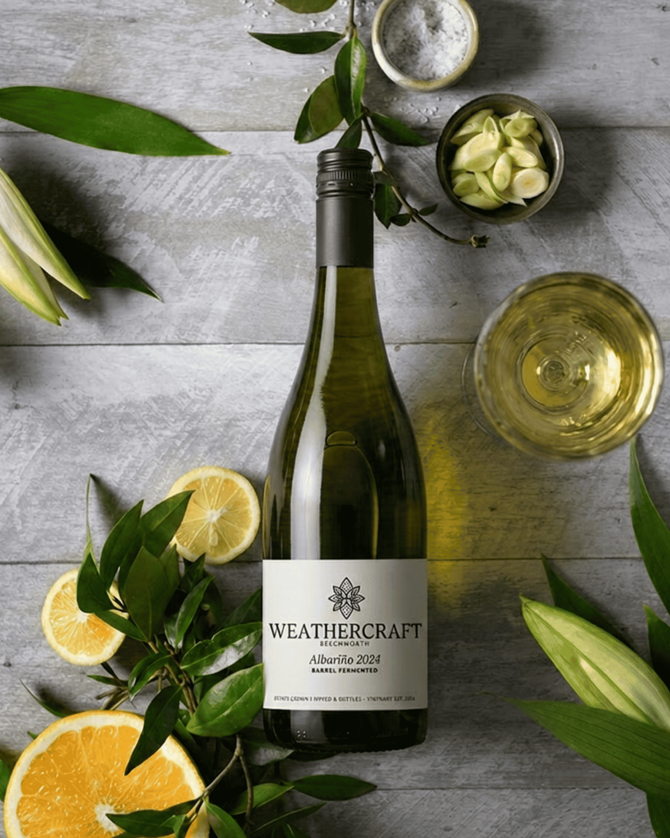 A bottle of Weathercraft Albariño 2024, styled in a flat-lay with lemon slices, garlic, olive leaves, and a glass of the pale yellow wine on a gray wood background.