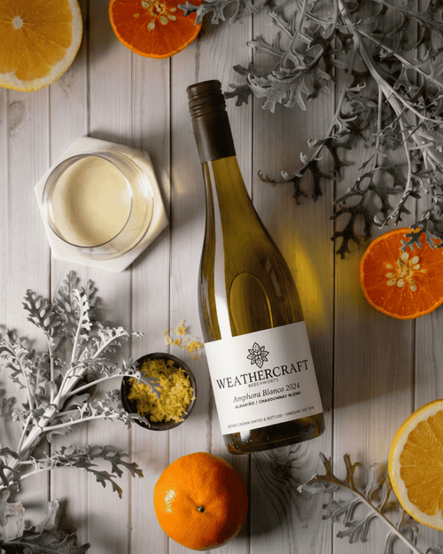 A bottle of Weathercraft Almond Blossom 2024 white wine is styled in a flat-lay composition on a light gray wooden surface, surrounded by orange slices, a glass of creamy white wine, and decorative silver foliage.