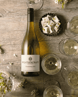 A bottle of Weathercraft Chardonnay 2024 white wine is displayed on a rustic wooden table alongside several stemmed wine glasses filled with the pale golden wine, a small bowl of white dessert cubes, and scattered white flowers.