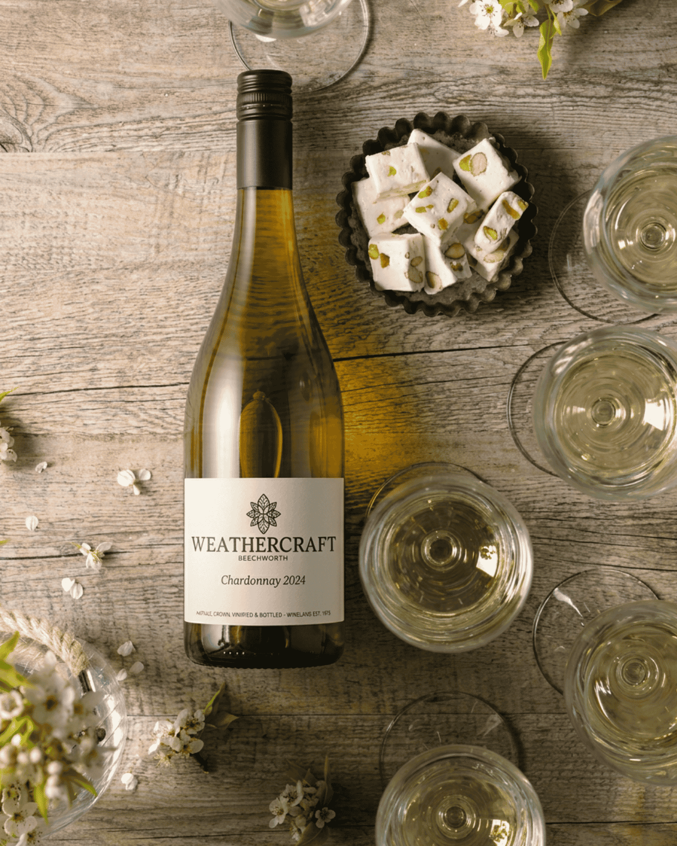 A bottle of Weathercraft Chardonnay 2024 white wine is displayed on a rustic wooden table alongside several stemmed wine glasses filled with the pale golden wine, a small bowl of white dessert cubes, and scattered white flowers.