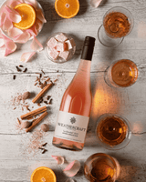 A bottle of Weathercraft La Ronche 2023 Rosé Grasse rosé wine is arranged on a light gray wooden surface with glasses of the pink wine, orange slices, cinnamon sticks, and pink petals.