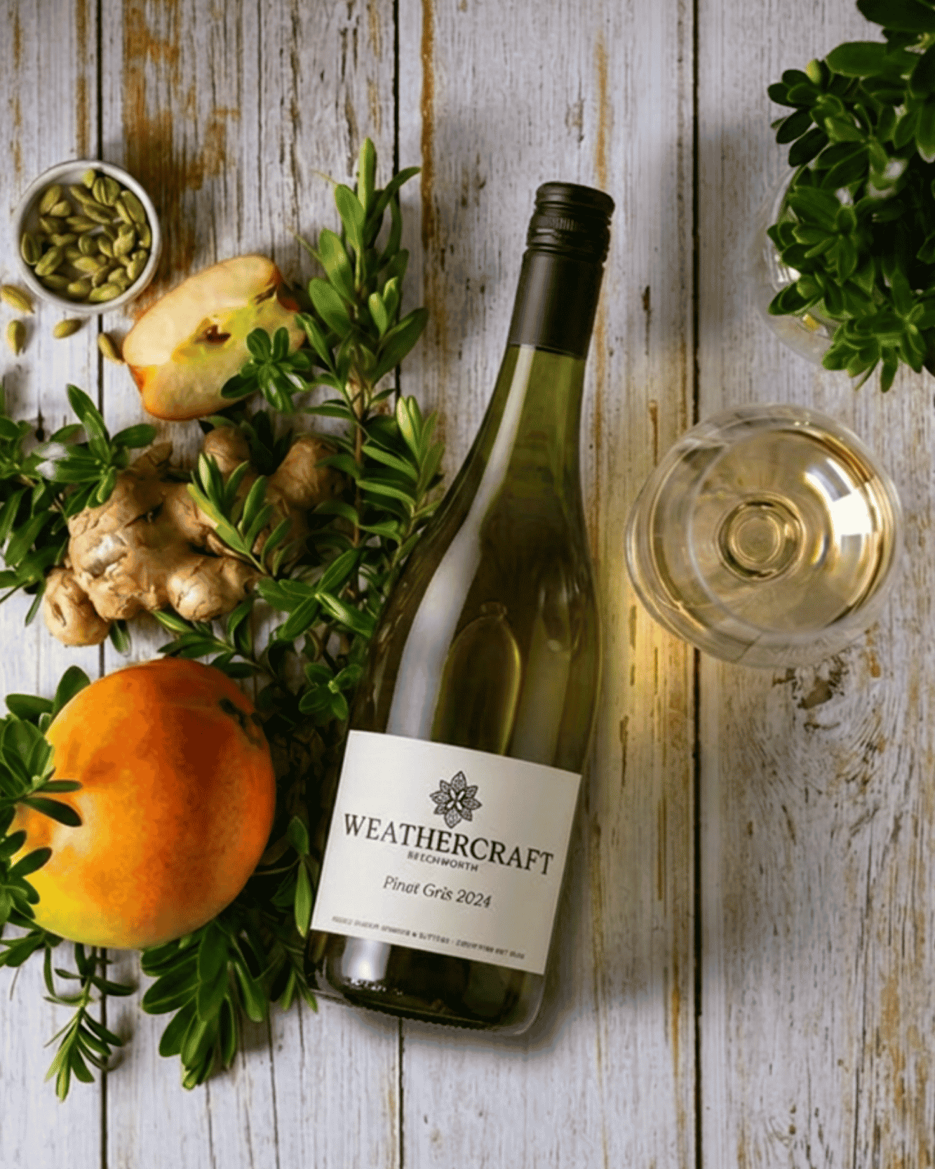 A bottle of Weathercraft Pinot Gris 2024 white wine with a glass of pale golden wine, styled on a rustic white wooden table with ginger, green herbs, and citrus slices.