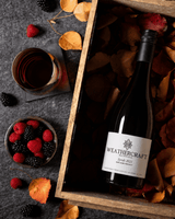 A bottle of Weathercraft Syrah 2021 wine nestled in a wooden gift box filled with autumn leaves, styled on a dark gray surface with fresh raspberries and a glass of red wine nearby.