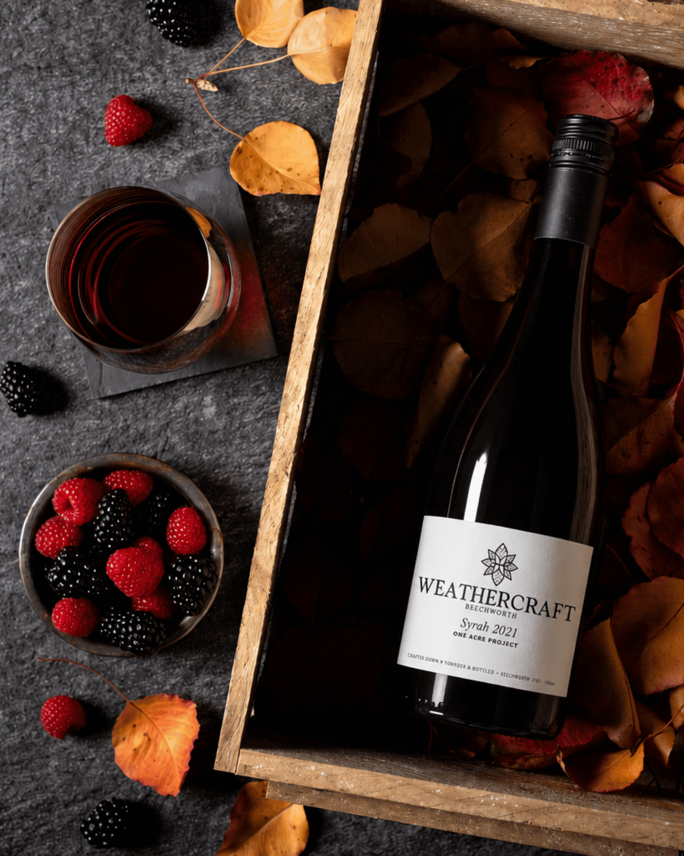 A bottle of Weathercraft Syrah 2021 wine nestled in a wooden gift box filled with autumn leaves, styled on a dark gray surface with fresh raspberries and a glass of red wine nearby.