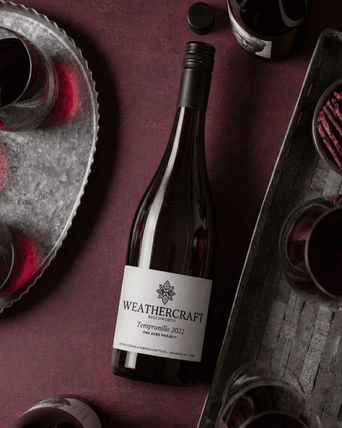 A bottle of Weathercraft La Tordera Tempranillo 2022 red wine, styled on a dark burgundy surface with galvanized trays and glasses of red wine.