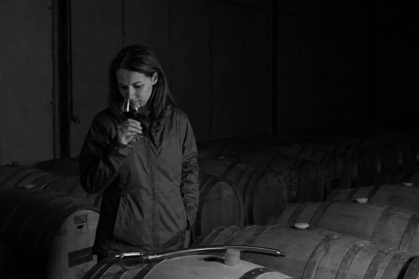 A woman in a dark jacket stands in a dim wine cellar or barrel room, holding a glass near her face, with large wooden barrels arranged around her.